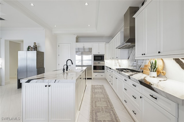 kitchen featuring backsplash, white cabinets, wall chimney range hood, built in appliances, and an island with sink