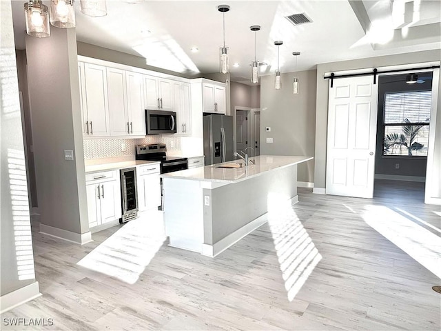 kitchen featuring appliances with stainless steel finishes, an island with sink, white cabinets, beverage cooler, and a barn door