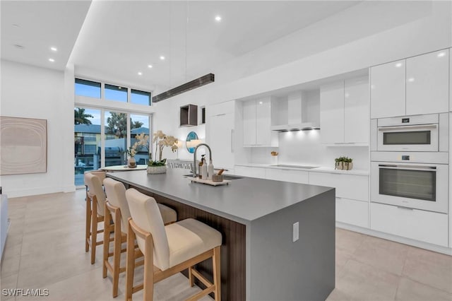 kitchen with wall chimney range hood, stovetop, a kitchen island with sink, a breakfast bar, and white cabinets