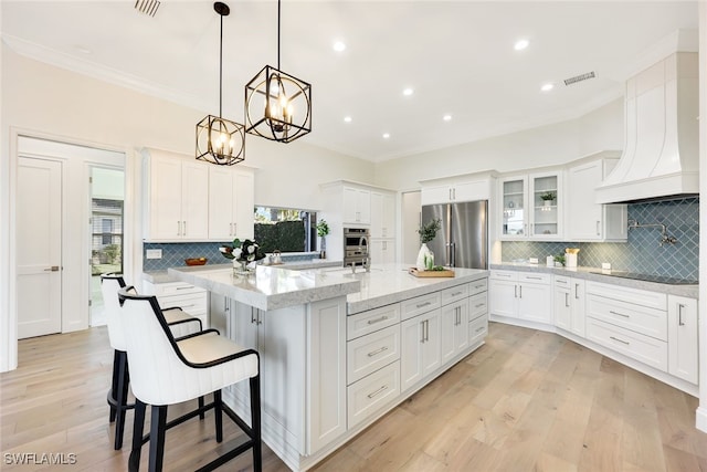 kitchen featuring a center island with sink, glass insert cabinets, appliances with stainless steel finishes, premium range hood, and white cabinetry