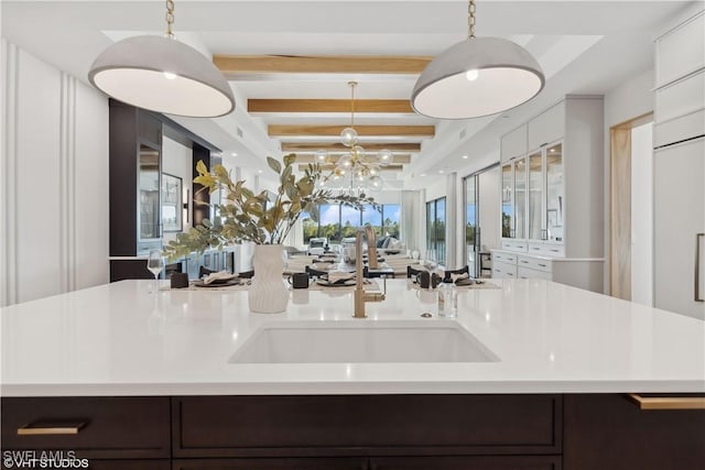 kitchen with pendant lighting, a kitchen island with sink, sink, dark brown cabinets, and beam ceiling