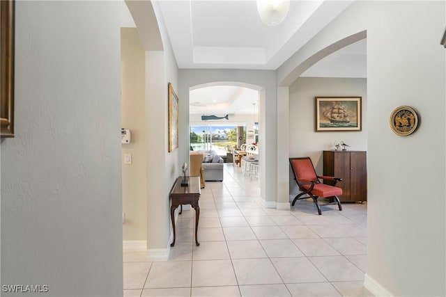 hallway with a tray ceiling, arched walkways, baseboards, and light tile patterned floors