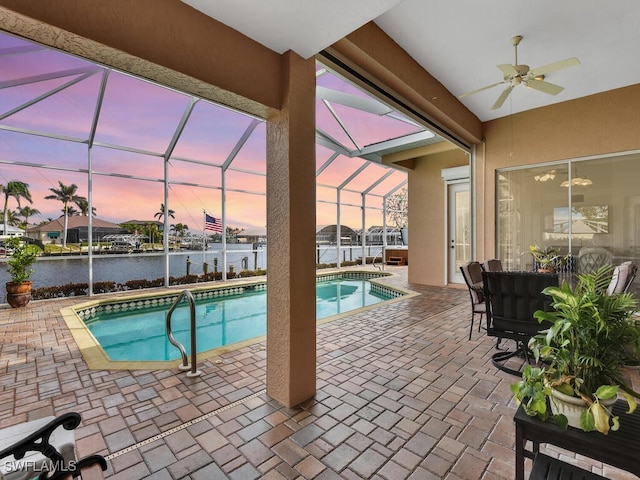 pool at dusk with ceiling fan, a patio area, a water view, and glass enclosure