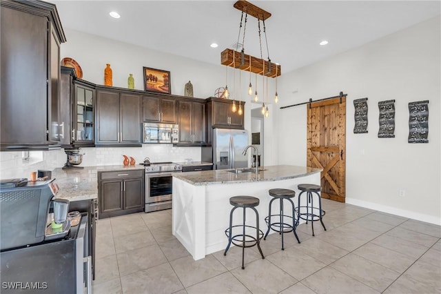 kitchen with a barn door, an island with sink, stainless steel appliances, hanging light fixtures, and sink