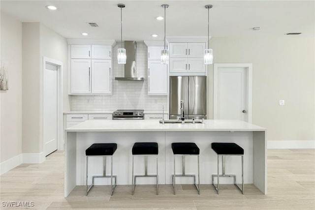 kitchen with backsplash, stainless steel appliances, a kitchen island with sink, wall chimney range hood, and white cabinetry