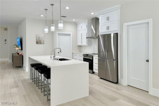 kitchen featuring sink, stainless steel appliances, wall chimney range hood, a center island with sink, and white cabinets