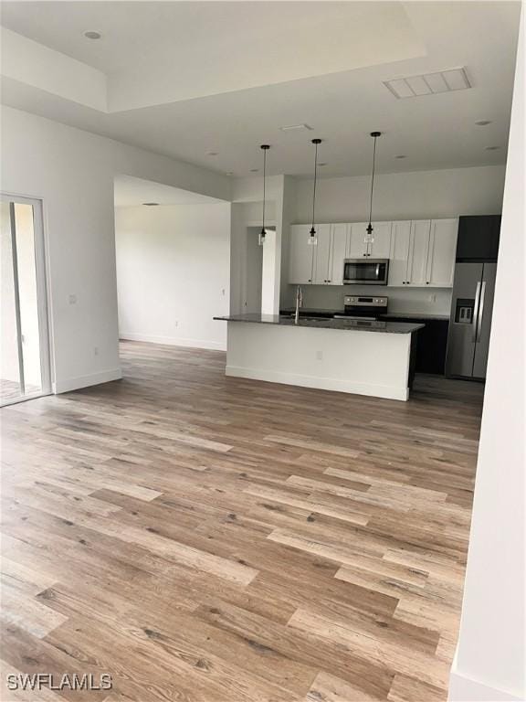 kitchen with light wood-type flooring, stainless steel appliances, white cabinets, hanging light fixtures, and an island with sink