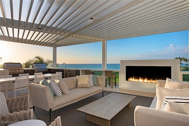 patio terrace at dusk with a view of the beach, a water view, a grill, an outdoor living space with a fireplace, and exterior kitchen