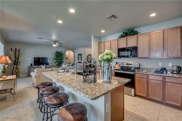 kitchen featuring ceiling fan, light stone countertops, stainless steel electric stove, a kitchen island with sink, and a kitchen bar