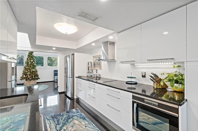 kitchen with black electric cooktop, stainless steel refrigerator, a raised ceiling, wall chimney range hood, and white cabinets