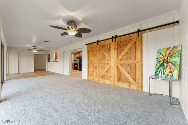 unfurnished living room with a barn door, ceiling fan, wooden walls, and light colored carpet