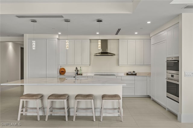 kitchen featuring a center island with sink, wall chimney range hood, hanging light fixtures, light tile patterned floors, and white cabinetry