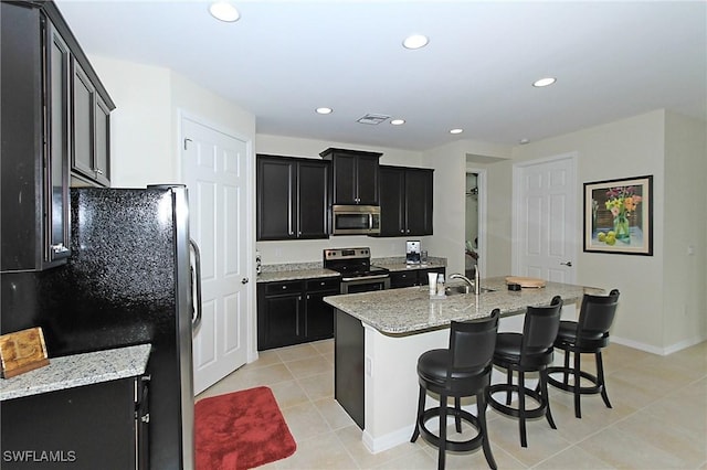 kitchen with stainless steel appliances, light stone counters, a kitchen bar, a kitchen island with sink, and light tile patterned floors