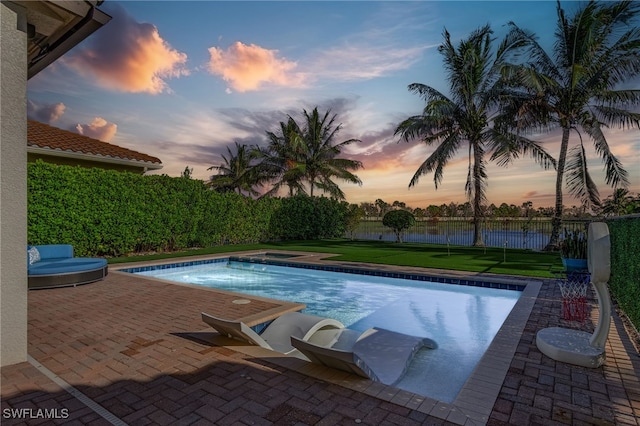 pool at dusk featuring a patio, a water view, and a lawn