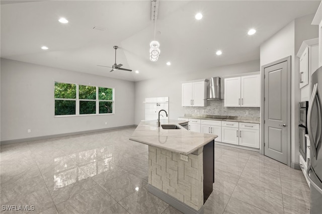 kitchen with white cabinets, light stone counters, wall chimney exhaust hood, and an island with sink