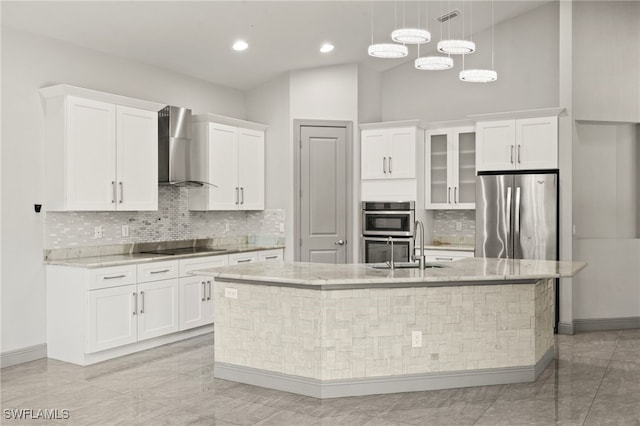 kitchen with appliances with stainless steel finishes, a kitchen island with sink, wall chimney range hood, white cabinets, and hanging light fixtures