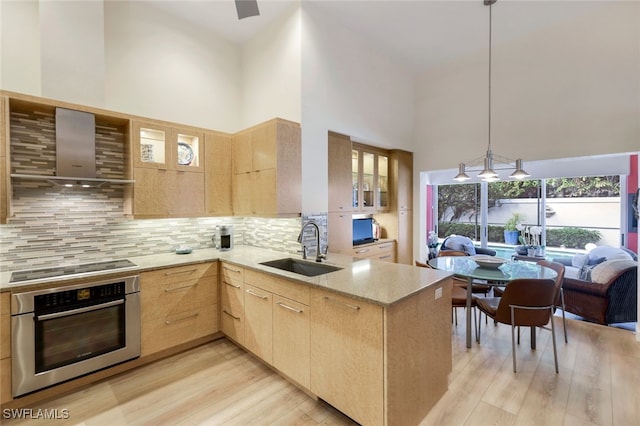 kitchen with kitchen peninsula, wall chimney exhaust hood, light brown cabinets, a high ceiling, and oven