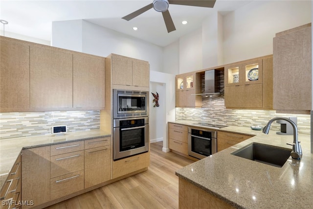 kitchen featuring appliances with stainless steel finishes, light stone counters, wall chimney exhaust hood, sink, and a high ceiling