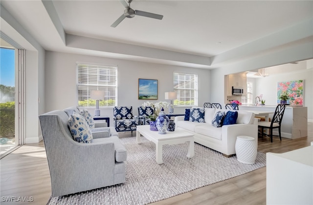 living area with a ceiling fan, a tray ceiling, baseboards, and light wood finished floors