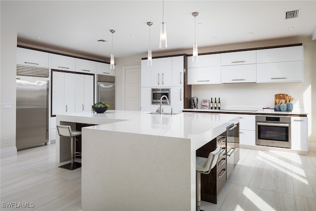 kitchen featuring built in appliances, white cabinetry, a center island with sink, and decorative light fixtures