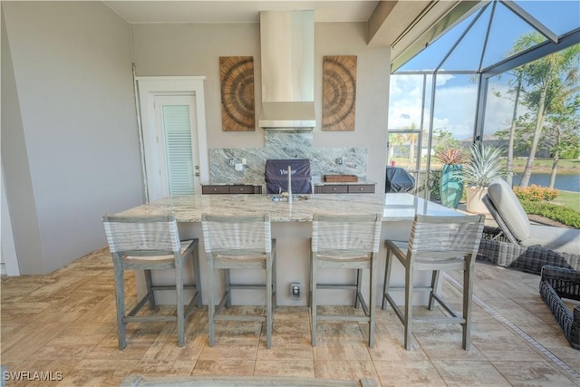 kitchen featuring a sunroom, wall chimney exhaust hood, light stone counters, a kitchen bar, and backsplash