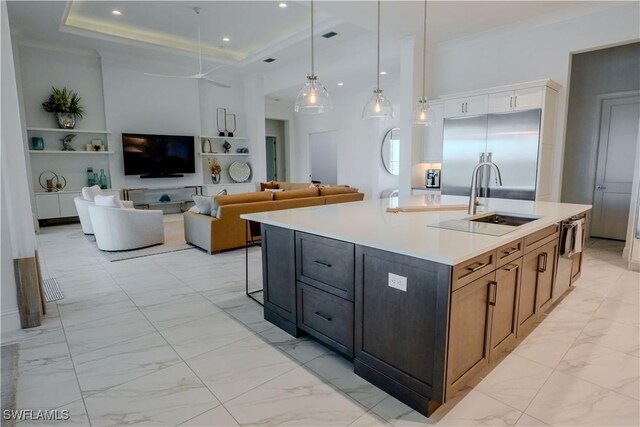 kitchen featuring a raised ceiling, sink, an island with sink, decorative light fixtures, and stainless steel built in refrigerator