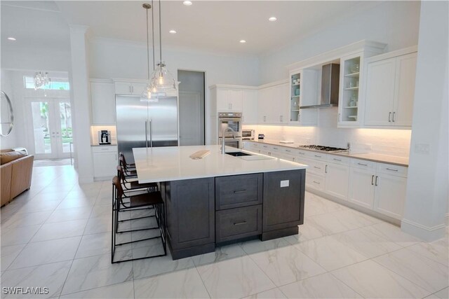 kitchen with stainless steel appliances, wall chimney range hood, pendant lighting, a kitchen island with sink, and white cabinets