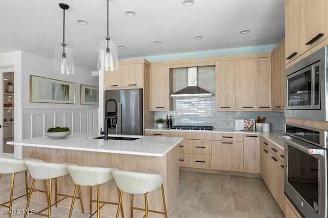 kitchen with appliances with stainless steel finishes, a center island with sink, light brown cabinetry, and wall chimney exhaust hood