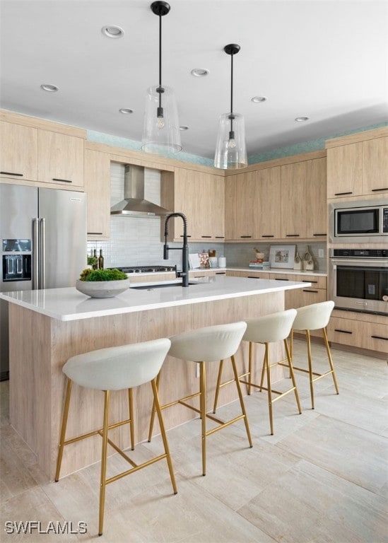 kitchen with wall chimney exhaust hood, light brown cabinetry, decorative light fixtures, a center island with sink, and appliances with stainless steel finishes