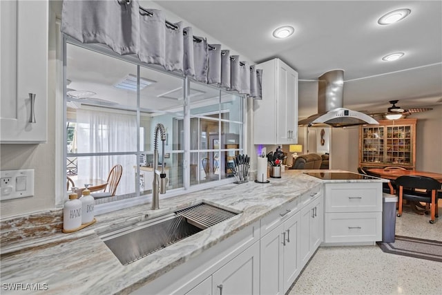 kitchen featuring sink, ceiling fan, island range hood, light stone counters, and white cabinetry