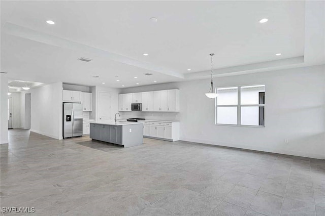 kitchen featuring appliances with stainless steel finishes, hanging light fixtures, a tray ceiling, an island with sink, and white cabinets