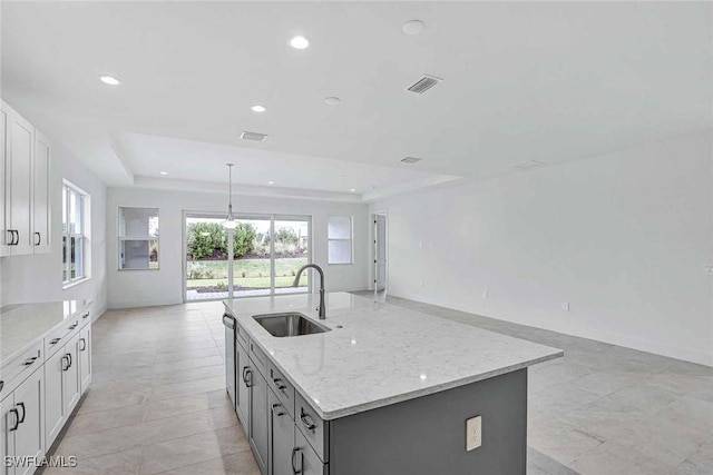 kitchen with sink, white cabinetry, a tray ceiling, light stone countertops, and an island with sink