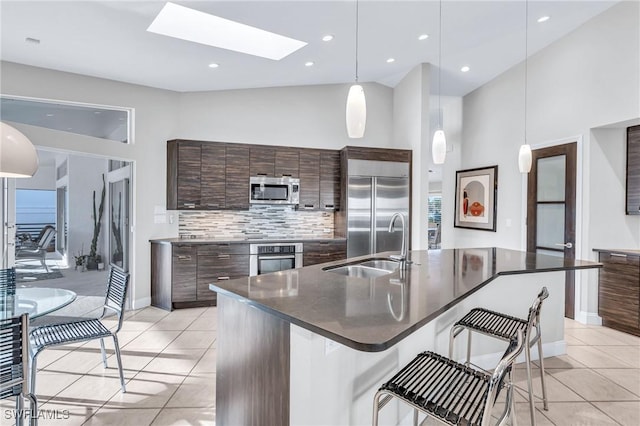 kitchen featuring appliances with stainless steel finishes, a skylight, dark brown cabinets, hanging light fixtures, and an island with sink