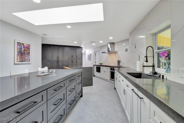 kitchen featuring a skylight, wall chimney exhaust hood, sink, white cabinetry, and oven