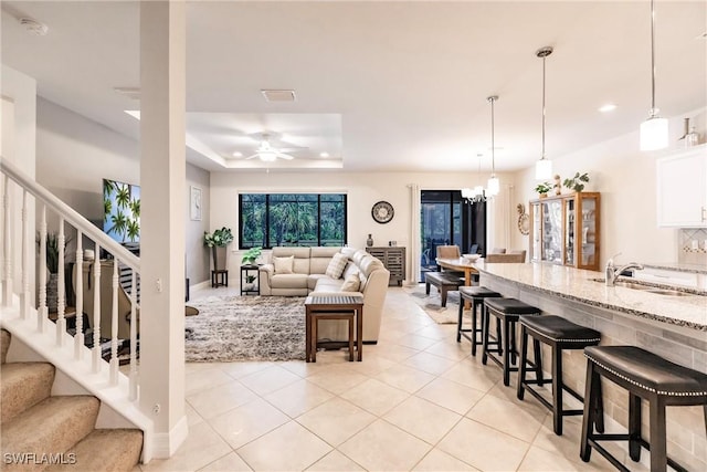 living room featuring light tile patterned floors, stairway, a tray ceiling, and ceiling fan with notable chandelier