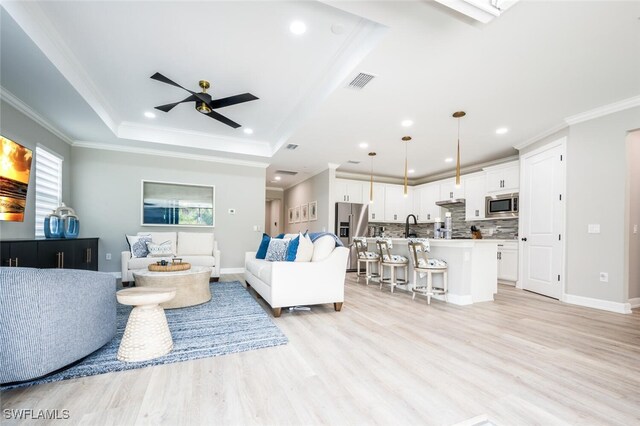 living room with ceiling fan, sink, light hardwood / wood-style flooring, a tray ceiling, and ornamental molding