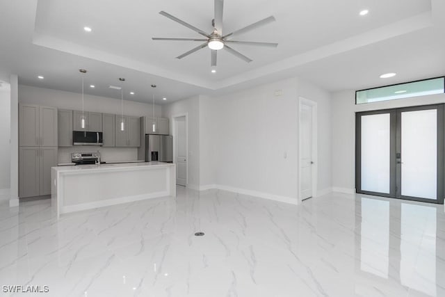 kitchen with gray cabinetry, stainless steel appliances, a raised ceiling, pendant lighting, and a center island with sink