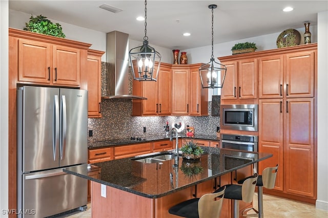 kitchen featuring sink, wall chimney exhaust hood, stainless steel appliances, pendant lighting, and a center island with sink