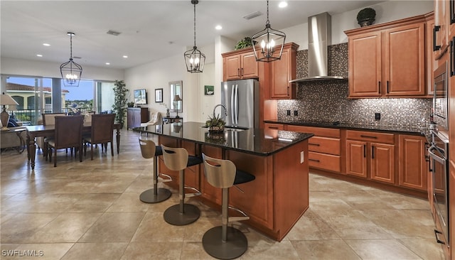 kitchen featuring a kitchen island with sink, wall chimney exhaust hood, decorative light fixtures, stainless steel refrigerator, and a chandelier