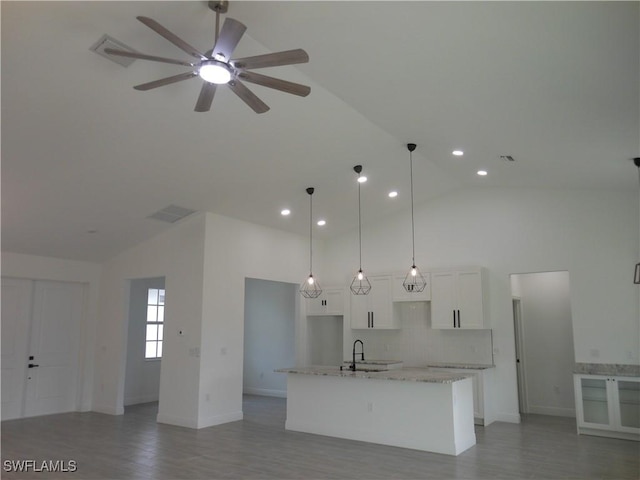 kitchen with light stone counters, a center island with sink, white cabinets, a sink, and high vaulted ceiling