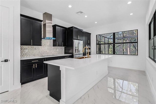 kitchen with sink, stainless steel appliances, wall chimney range hood, tasteful backsplash, and an island with sink