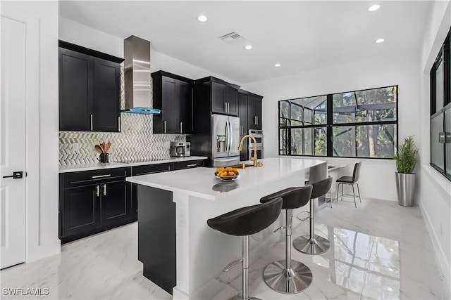 kitchen with a center island with sink, stainless steel refrigerator with ice dispenser, wall chimney exhaust hood, decorative backsplash, and a breakfast bar area