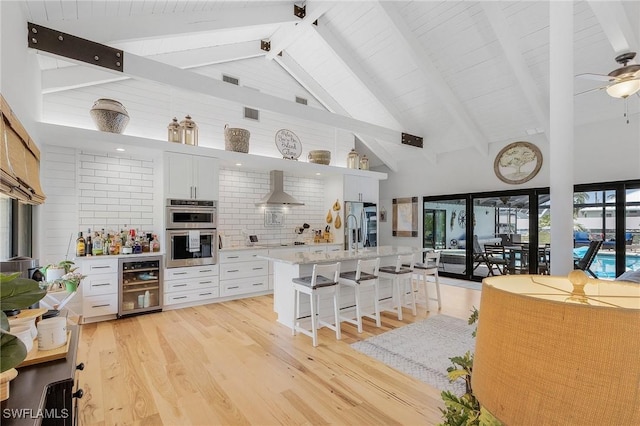 kitchen featuring high vaulted ceiling, wall chimney exhaust hood, white cabinetry, and wine cooler