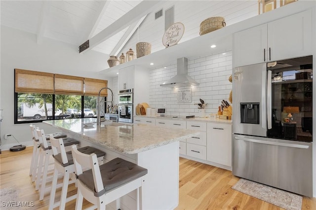 kitchen with beam ceiling, wall chimney range hood, a center island with sink, white cabinets, and appliances with stainless steel finishes