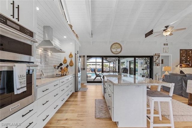 kitchen with a kitchen island with sink, white cabinets, wall chimney range hood, light stone countertops, and a kitchen bar