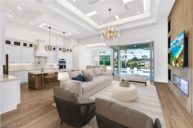 living room with a glass covered fireplace, a towering ceiling, light wood-style flooring, an inviting chandelier, and a tray ceiling