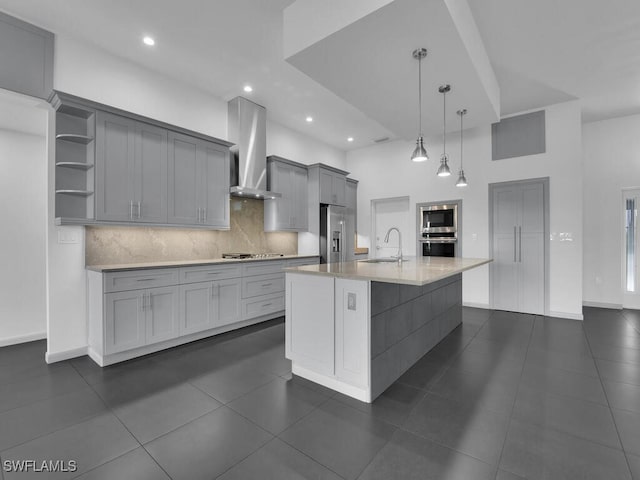 kitchen with wall chimney exhaust hood, sink, hanging light fixtures, gray cabinets, and an island with sink