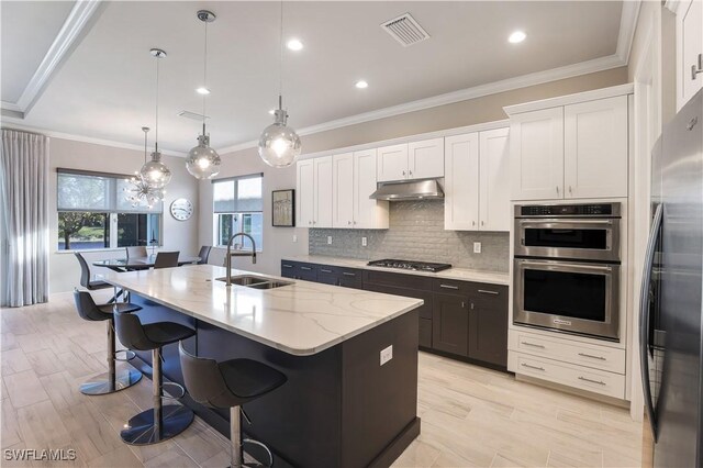 kitchen with stainless steel appliances, a kitchen island with sink, sink, white cabinetry, and hanging light fixtures