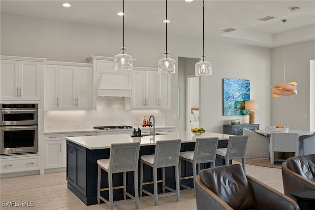 kitchen featuring premium range hood, a kitchen island with sink, white cabinets, and stainless steel double oven