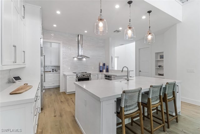 kitchen with a large island with sink, white cabinets, wall chimney range hood, sink, and hanging light fixtures